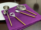 Set of four sleek bartending tools with gold handles and silver heads, including a jigger, heart-shaped bottle opener, cocktail strainer, and spoon, arranged on a purple cloth.