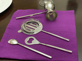 Set of four silver-colored bar tools including a jigger, a spring strainer, a bottle opener with a heart-shaped hole, and a stirring spoon, arranged on a vibrant purple cloth.