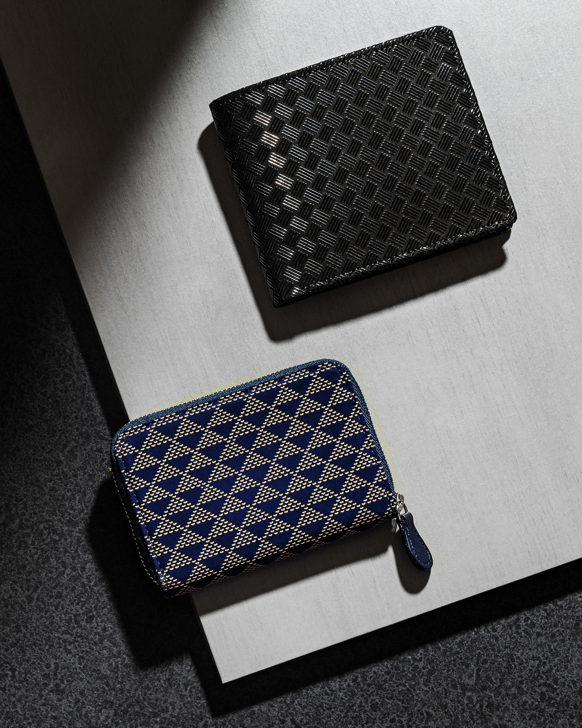 Two compact wallets on a layered surface; top is a black bi-fold with a checkerboard texture, bottom is a navy zip-around with beige geometric triangle pattern.