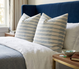 Two square pillows with horizontal blue and natural stripes on a navy blue upholstered headboard bed, alongside white bedding and a bedside table with a bowl and book.