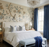 Bedroom with large bed featuring white linens and three blue-and-white patterned pillows, botanical bird wallpaper, dark indigo curtains, and a blue bench with a draped blanket.