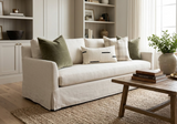 Neutral-toned living room featuring a beige sofa with five pillows including an ivory Dashes Print Mudcloth pillow, green velvet cushions, and plaid accents, facing a wooden coffee table on a woven rug.