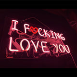 Pink neon sign displaying the phrase "I F❤cking Love You" with a red heart replacing the letter O, mounted on a transparent backed panel in a dark setting.