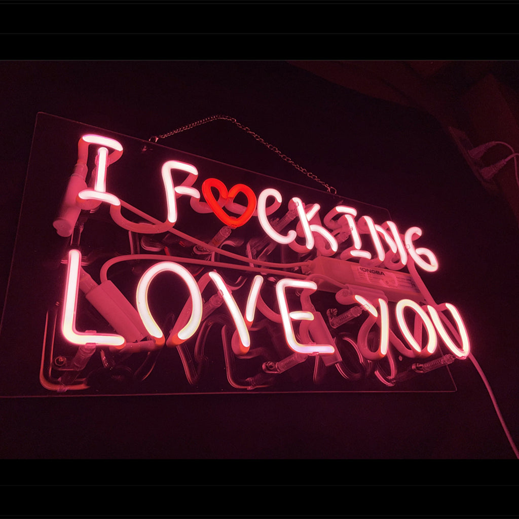 Pink neon sign displaying the phrase "I F❤cking Love You" with a red heart replacing the letter O, mounted on a transparent backed panel in a dark setting.