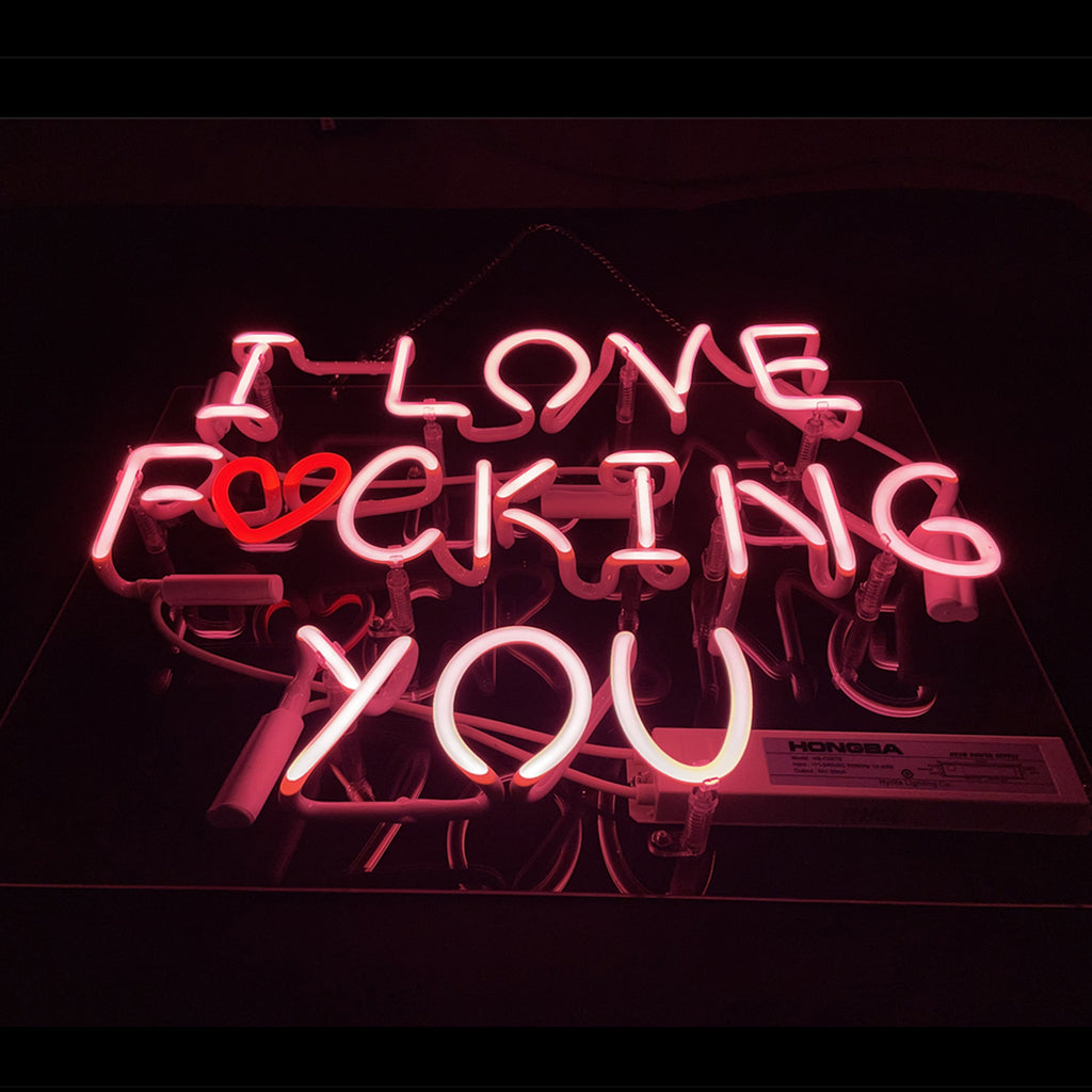 Pink neon sign displaying the phrase 'I LOVE FUCKING YOU' with a red heart shape replacing the letter 'U' in 'FUCKING' on a dark background.