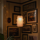 White wavy pendant light glowing warmly, hanging in a corner surrounded by multiple framed artworks and photos in black frames on beige walls.