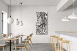 Vertical abstract artwork with black ink swirls and textured gray background hanging on white brick wall in modern café with wooden chairs and stools.