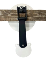 White circular metal shooting target with a smaller swinging target attached below, mounted on a horizontal weathered wooden beam with black metal bracket hardware.
