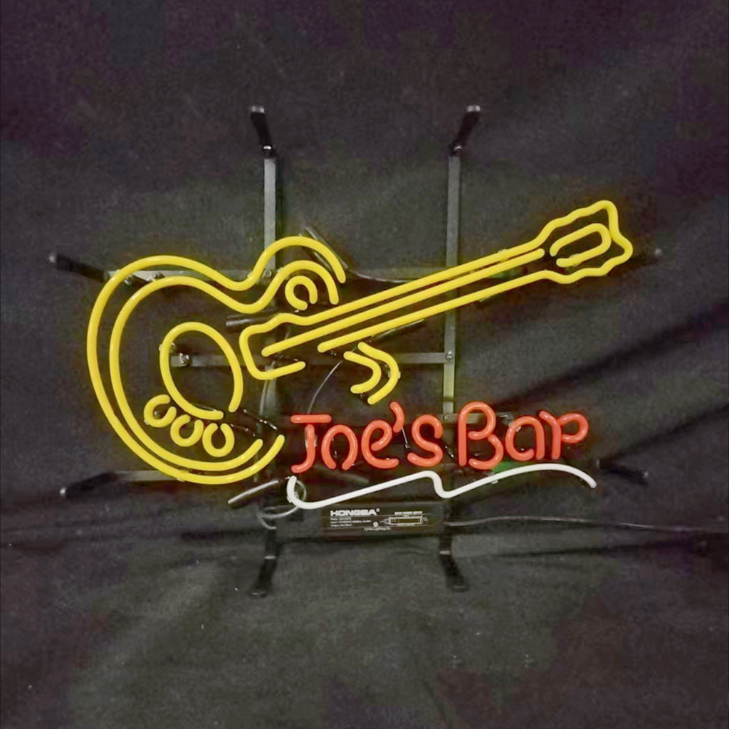 Yellow neon light shaped like an electric guitar with red neon text below spelling 'Joe's Bar' and a white neon underline, mounted on a metal stand against a black background.