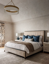 Bedroom with a beige upholstered bed adorned with seven pillows including blue pillows with a wavy pattern, flanked by nightstands and table lamps under a circular chandelier.