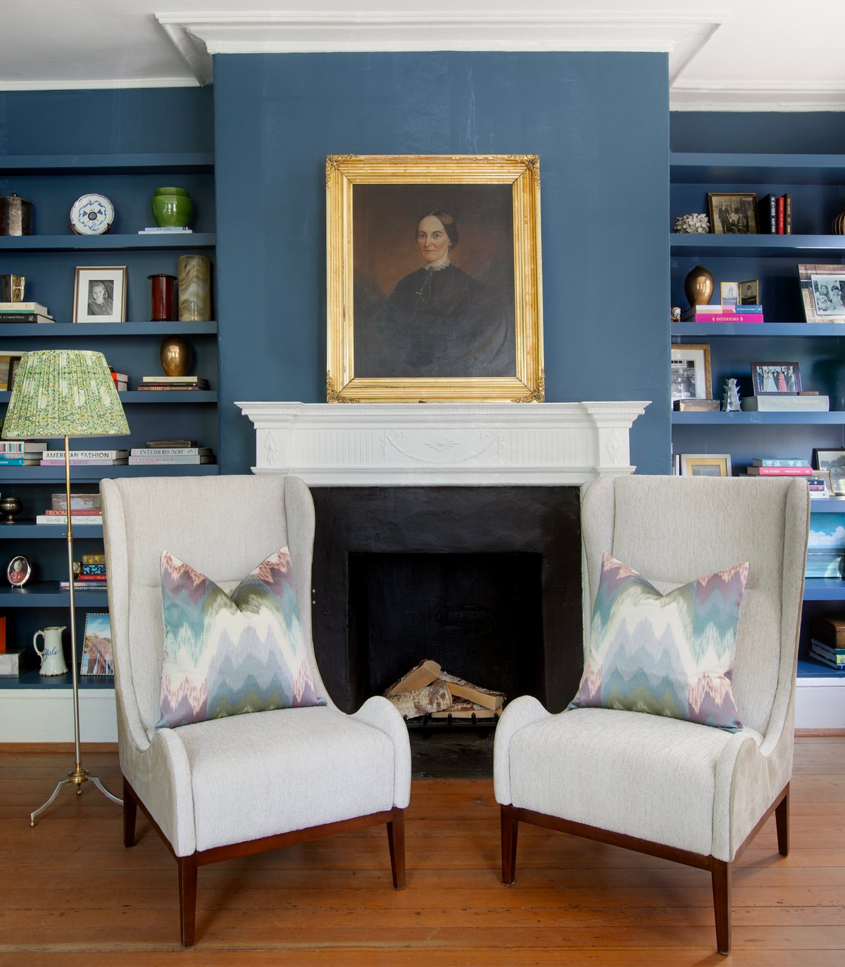 Two cream wingback chairs with chevron-patterned pastel pillows face a white fireplace set in a blue wall with built-in shelves holding books and decor.
