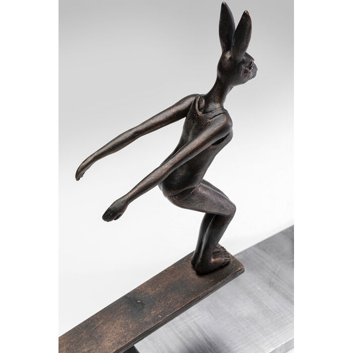 Bronze-colored figurine of a humanoid rabbit with elongated ears balancing on a narrow, rectangular base, shown in a side view against a light background.