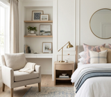 Beige armchair with a folded cream blanket in a neutral bedroom featuring a side table with brass lamp, bed with pink and blue paisley patterned pillows, and a round mirror above.