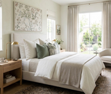 Neutral-toned bedroom with a beige upholstered bed, white and sage green pillows, wooden nightstand with gold lamp, and large floral wall art above bed.