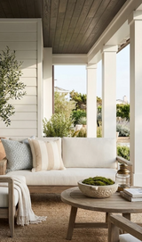Outdoor seating area with beige cushioned sofa featuring Ketley Stripe Pillow Cover in natural tones and blue patterned pillow, next to wood round coffee table with a moss bowl and lantern.