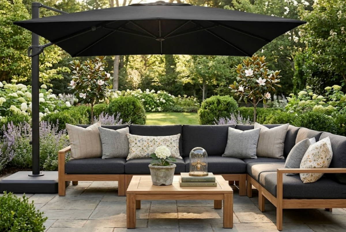 Outdoor seating area with a wooden-framed L-shaped sofa featuring dark cushions and neutral patterned pillows, shaded by a large black cantilever umbrella over a light stone patio.