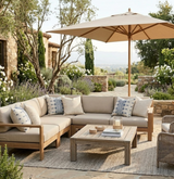 Outdoor patio with beige sectional sofa featuring cream and indigo-patterned pillows, a wooden coffee table, and a large tan umbrella against a stone wall and greenery.