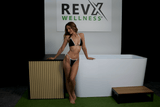 Woman in black and white bikini leaning on a vertical slatted wooden panel beside a white acrylic cold plunge tub and small wooden step, under a RevX Wellness sign.