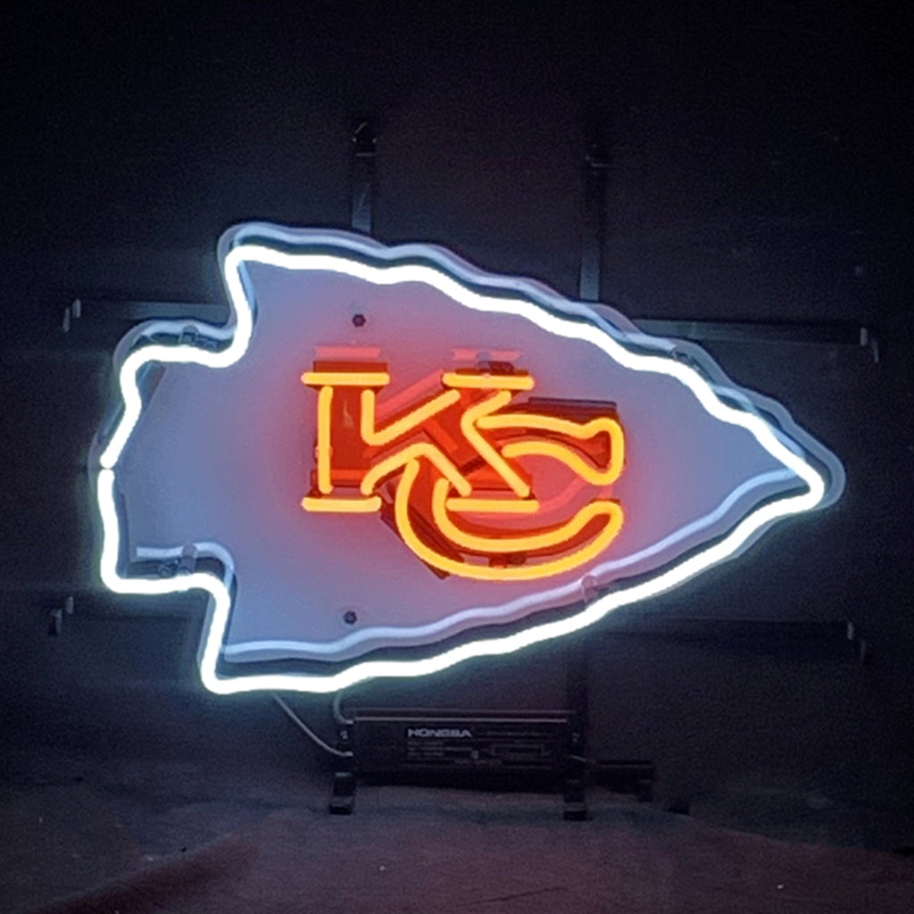 Neon sign shaped like an arrowhead with white outline and red interlocking KC letters inside, glowing against a dark background.