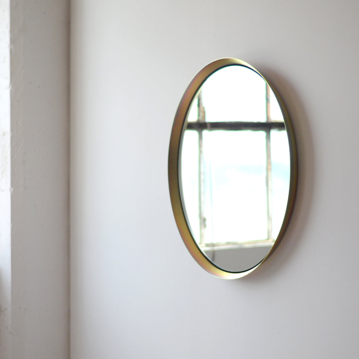 Round wall mirror with a thin gold frame reflecting a window with grid lines, mounted on a plain light-colored wall.