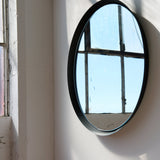 Round wall mirror with thin black frame reflecting an industrial-style window and blue sky, mounted on a light-colored wall near a window.
