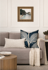 Square pillow with large white leaf pattern on mineral blue background on a gray sofa, next to a long light beige textured pillow and beige throw.