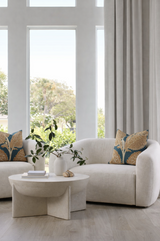 Curved cream sofa with pillows featuring ochre floral patterns on mineral blue background, beside a round beige coffee table with white vases holding green leafy branches.