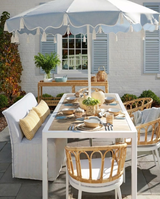 Outdoor dining setup with white metal table and rattan-accented chairs, featuring beige tableware, yellow patterned pillows, and a white scalloped umbrella.