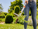 Person using a metal weed puller tool to remove a dandelion with roots from a garden lawn, against a backdrop of green bushes and plants in sunlight.