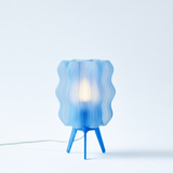 Blue wavy translucent lamp shade on a three-legged blue base with a glowing light bulb inside, shown against a plain white background.