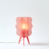 Red table lamp with wavy slatted shade and three tripod legs, softly glowing against a white background