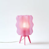 Pink translucent lamp with wavy vertical slats around the bulb and three angled legs, glowing softly against a white background.