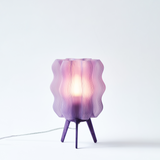 Purple wavy-patterned lamp shade on a three-legged purple base, glowing softly from an internal light source against a white background.