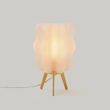 Small table lamp with a wavy white translucent shade on a yellow three-legged base, glowing softly against a plain light gray background.