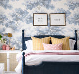 Bedroom with a dark blue wooden bedframe, featuring saffron yellow and pink patterned pillows against blue and white leafy wallpaper and framed wall art.