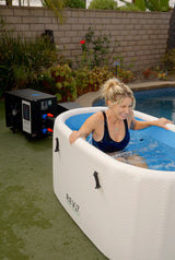 Smiling woman in a black swimsuit relaxing in a white inflatable hot tub with blue interior on a green outdoor surface near a brick wall and garden plants.