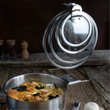 Stainless steel pot with cooked food in front of a metal and chrome circular lid holder holding three glass lids with black handles, set on rustic wooden surface.