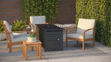 Outdoor seating area with three wooden armchairs upholstered in light fabric around a black slatted rectangular fire pit table, next to a small wooden side table with plant, surrounded by greenery and brick wall.