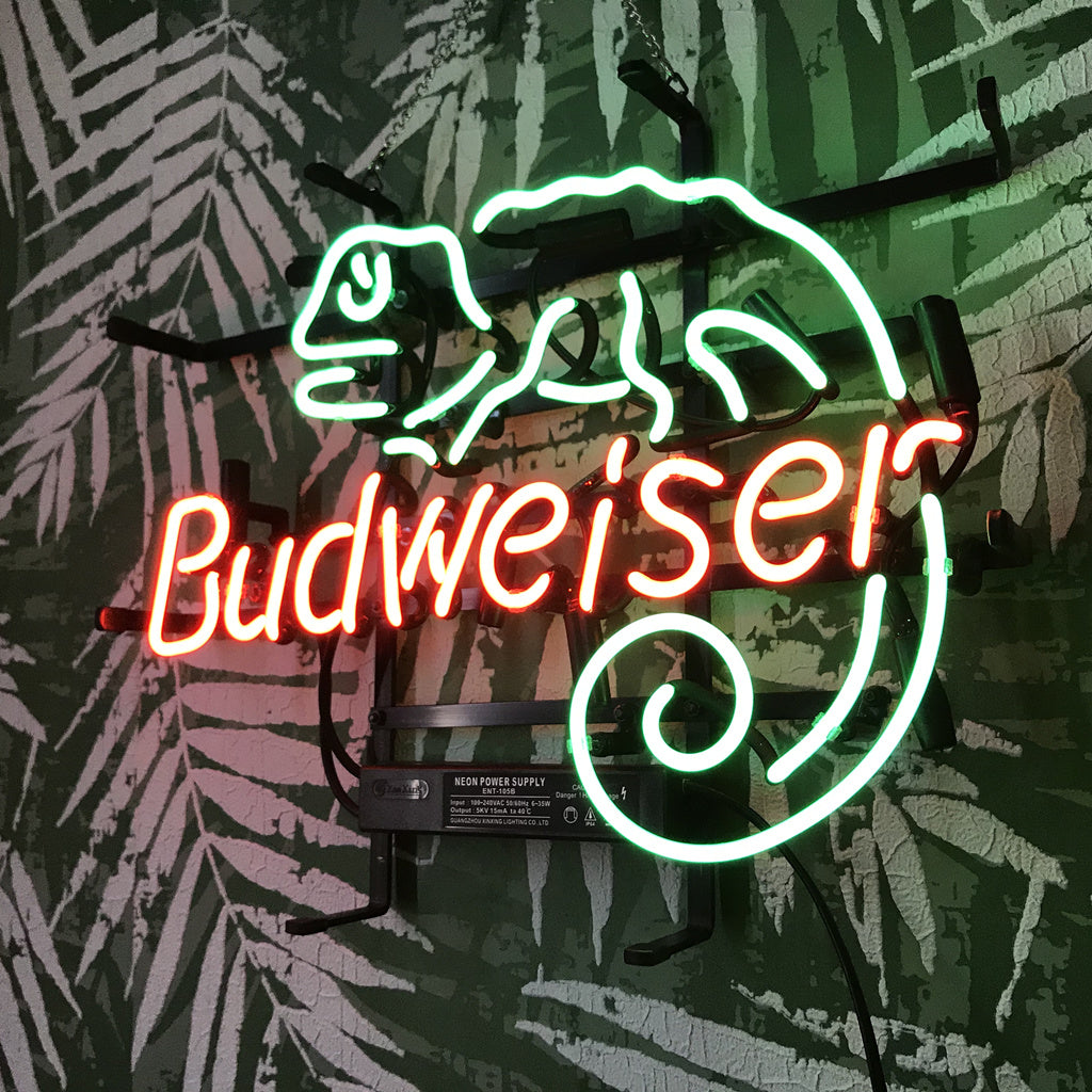 Neon light sign with a green lizard outline curling its tail above red 'Budweiser' text, mounted on a dark green wall with white fern leaf patterns.