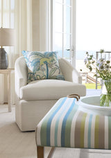 Off-white upholstered armchair with a decorative pillow featuring blue and green tropical leaf patterns, next to a striped ottoman in blue, green, and white shades.