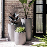 Group of three smooth, cylindrical gray planters holding different plants, set on a brick floor against a brick wall near a black-framed window.