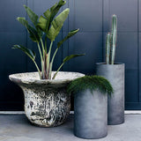 Three modern planters against a dark blue paneled wall: a wide, textured beige and black planter with tall green-leafed plant, a short gray cylindrical planter with trailing greenery, and a taller gray cylindrical planter with a s