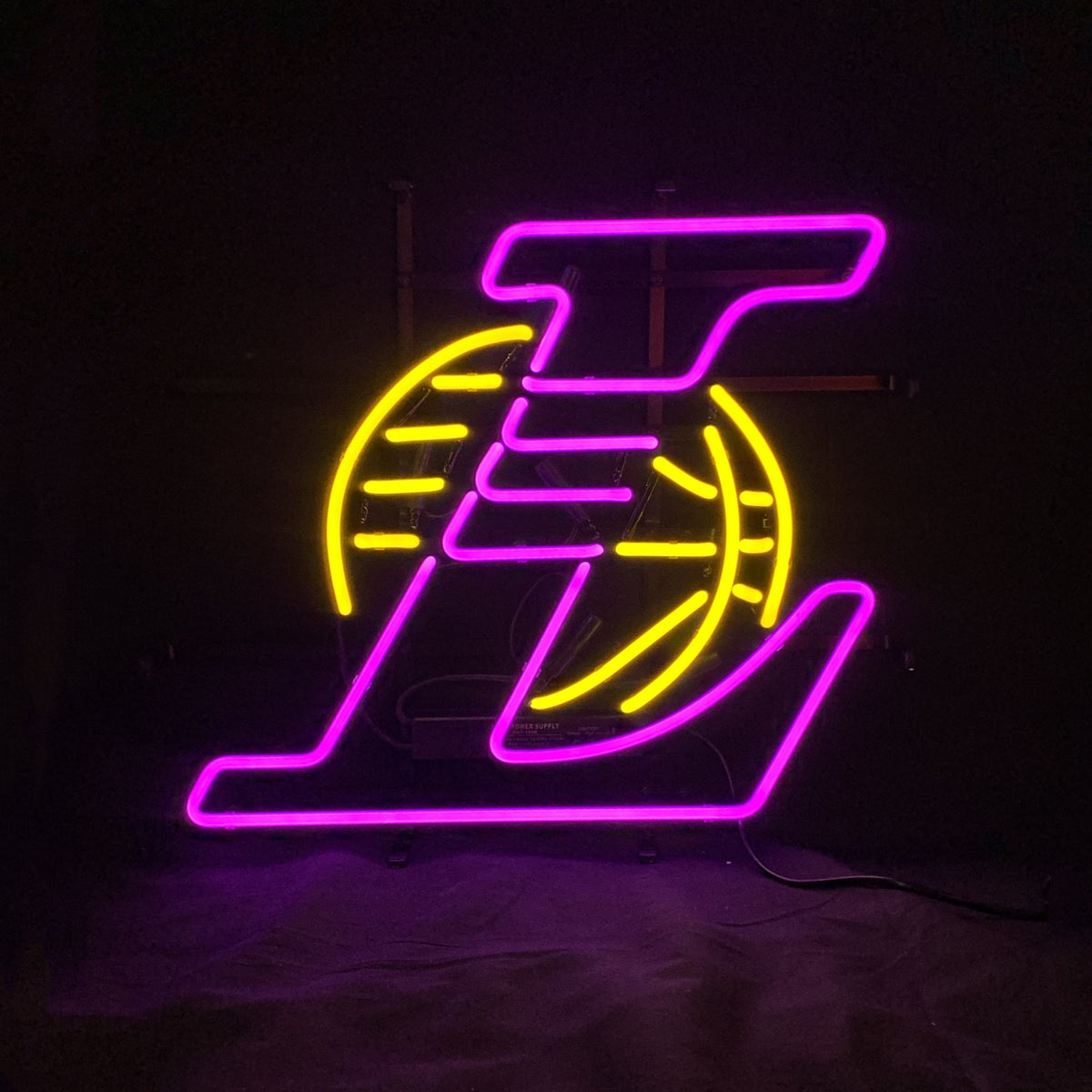 Purple neon Los Angeles Lakers 'L' logo overlaying a yellow neon basketball outline on a black background, glowing softly in the dark.