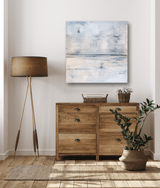 Square canvas print with abstract light blue and white brushstrokes above wooden dresser, accompanied by a tripod floor lamp and potted green plant nearby.
