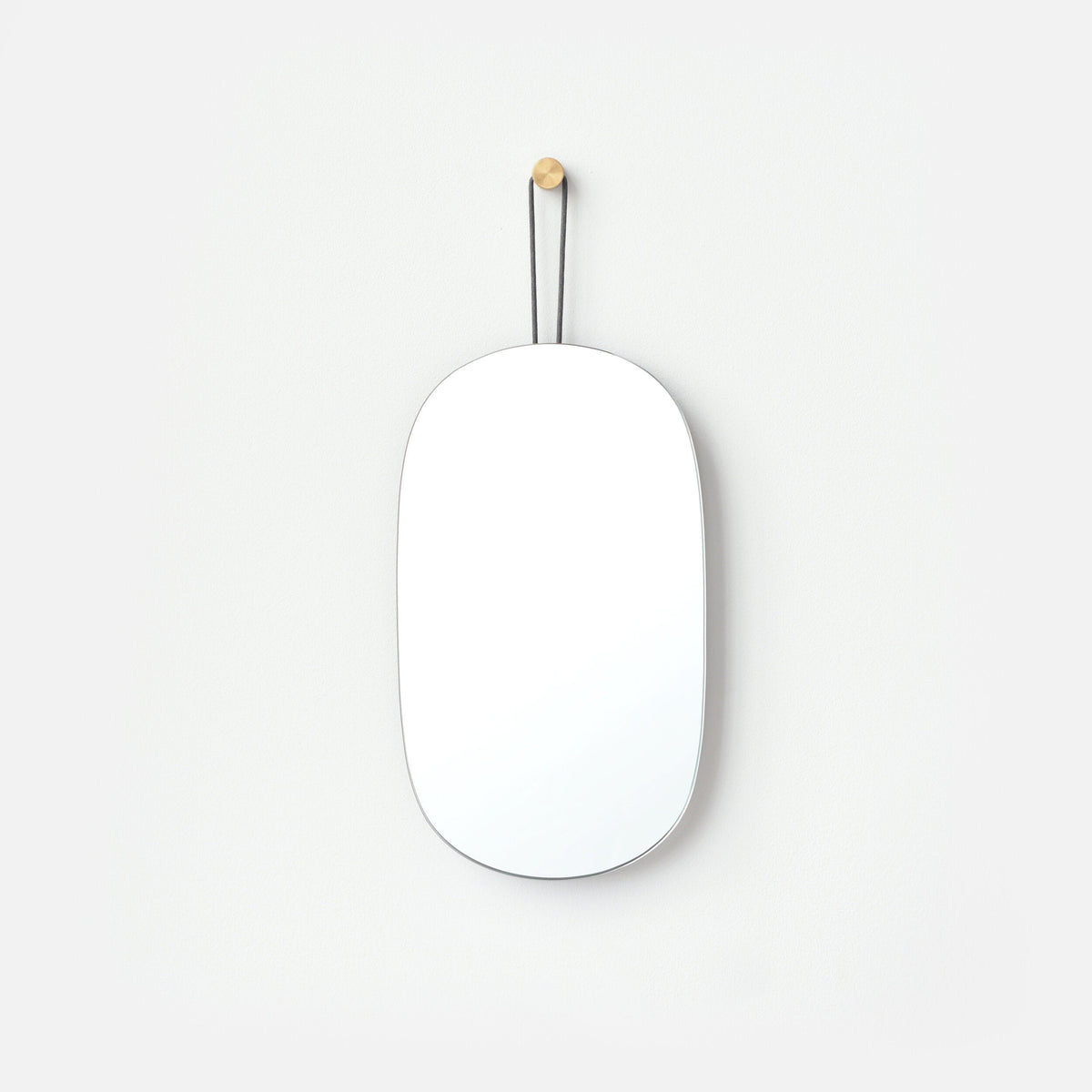 Minimalist oval mirror with rounded edges hanging on a white wall by a thin black strap attached to a small wooden peg.