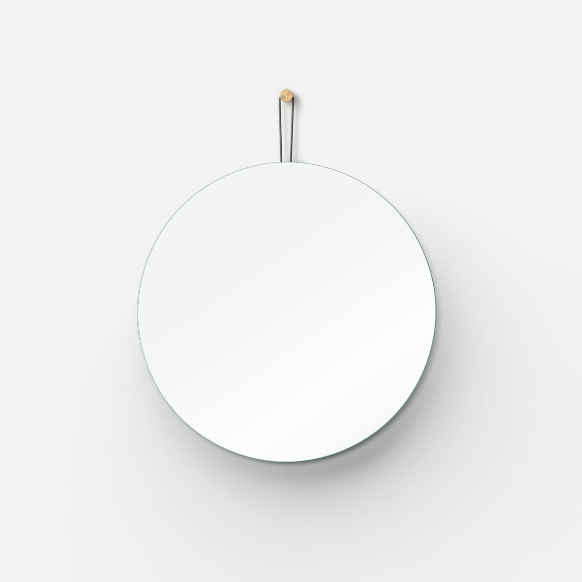 Round wall mirror with a thin, frameless edge, hanging from a leather strap attached to a wooden peg on a white wall.