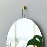 Round wall mirror with thin black frame hung by two black straps attached to a gold circular wall mount, reflecting a blurred window and a green leaf.