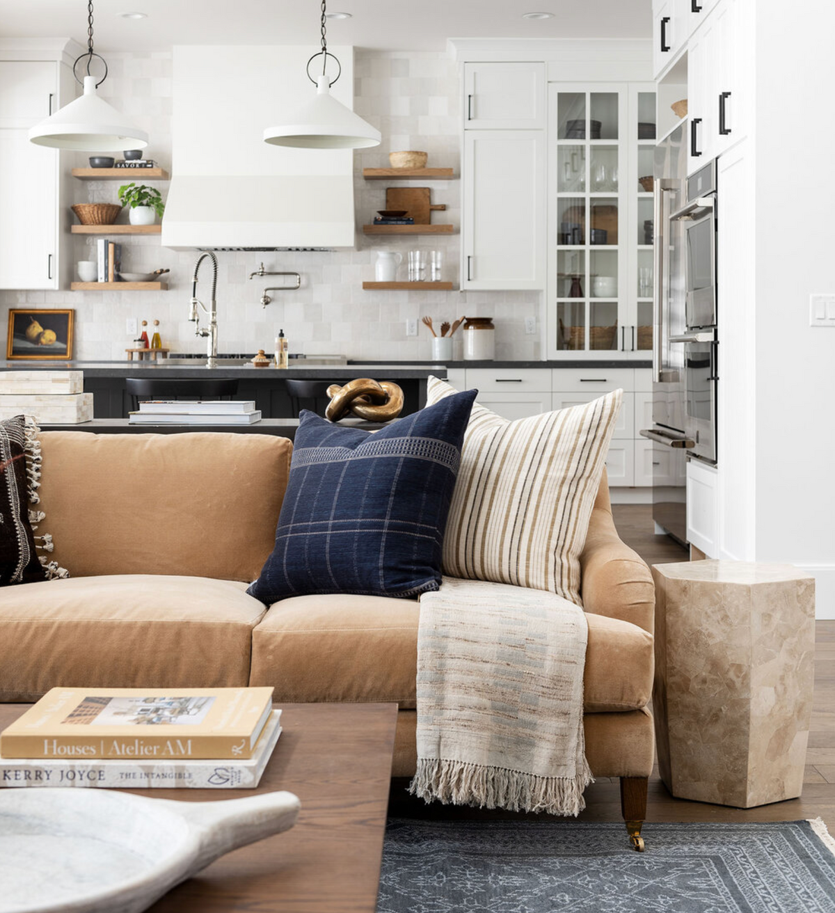 Tan sofa with a blue plaid pillow and a striped pillow with a fringe throw draped over the arm, set in front of a white kitchen with open shelves and pendant lights.