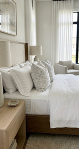 Bedroom with bed featuring Marbella Pillow Covers in smoke pattern, white bedding, beige headboard, wooden nightstand, and sheer white curtains on tall windows.