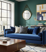 Indigo blue sofa with cylindrical armrests, adorned with patterned and solid pillows, set against deep green walls with geometric mirror and abstract gold-accented artwork.